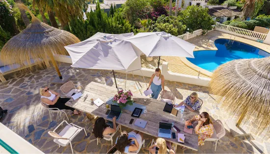 Overhead shot of Kate and group meeting at a large round outdoor table, shaded by umbrellas.