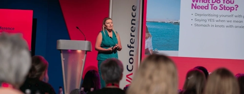 Kate Kurdziej speaking on stage with a lectern on her left and slide show on her right.
