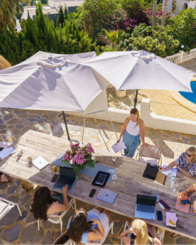 Overhead shot of Kate and group meeting at a large round outdoor table, shaded by umbrellas.